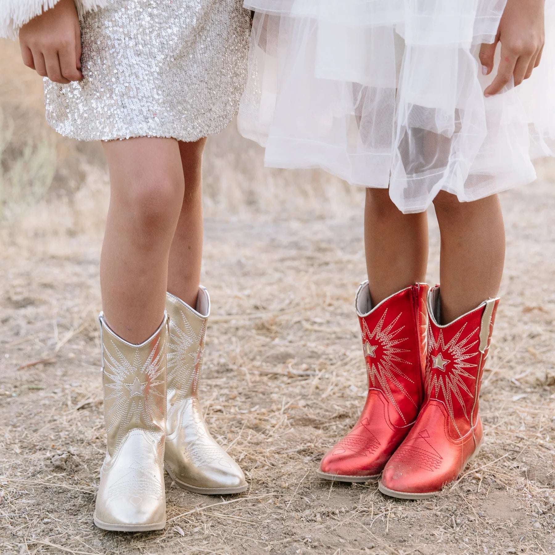 Two pairs of boots, one gold and one red, worn by children in white dresses on a natural background.