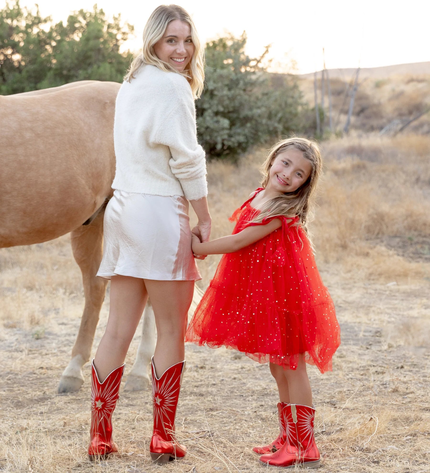 Woman and young girl in red cowboy boots standing next to a horse in a natural setting.