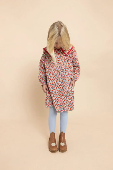 A young girl with blonde hair is wearing a long-sleeved dress with a red, white, and blue floral pattern, light blue leggings, and brown boots, standing against a solid beige background.