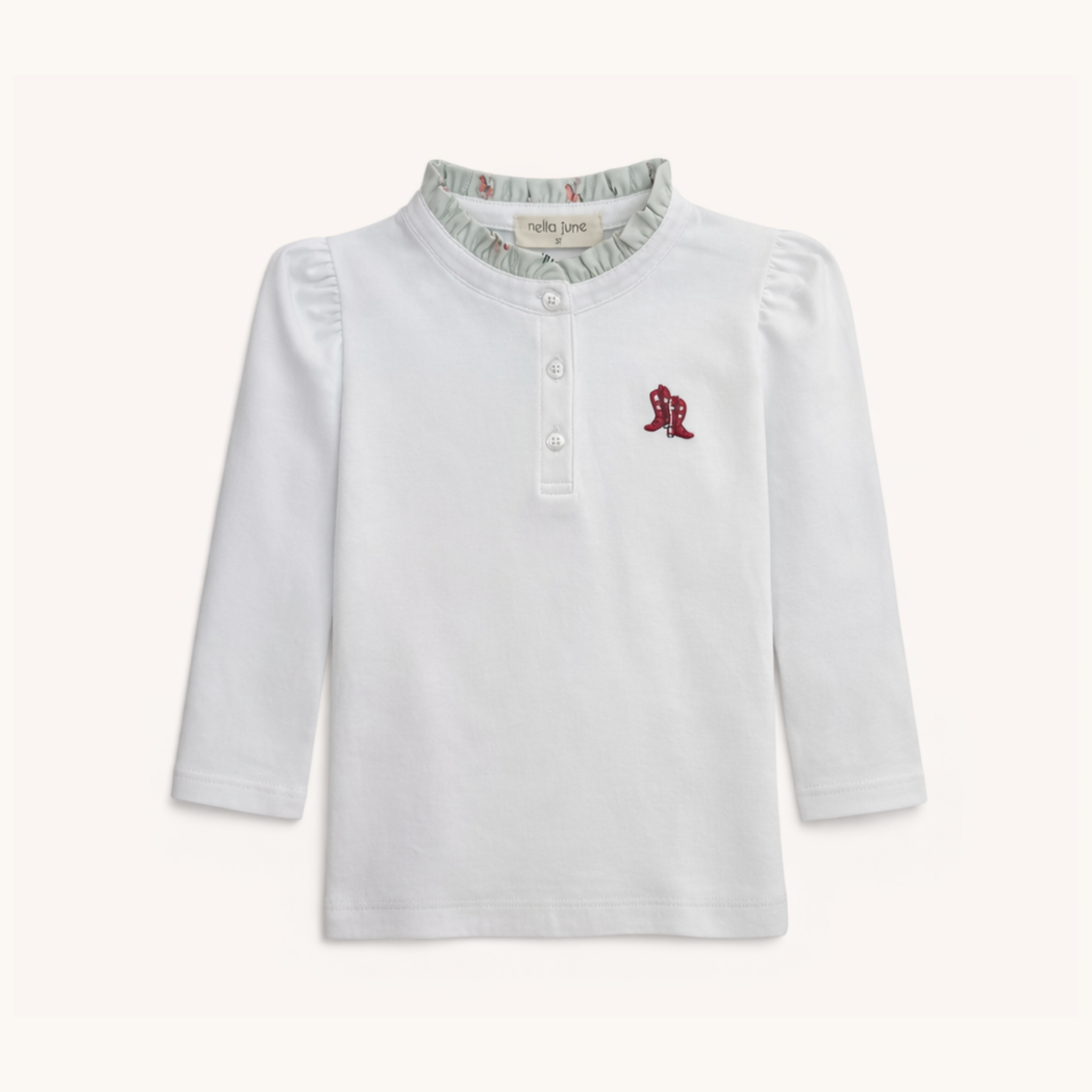 A white long-sleeve baby henley top by Nella Jane, featuring a ruffled collar with a light green rodeo pattern, a three-button placket, and a small embroidered red boot on the left chest.