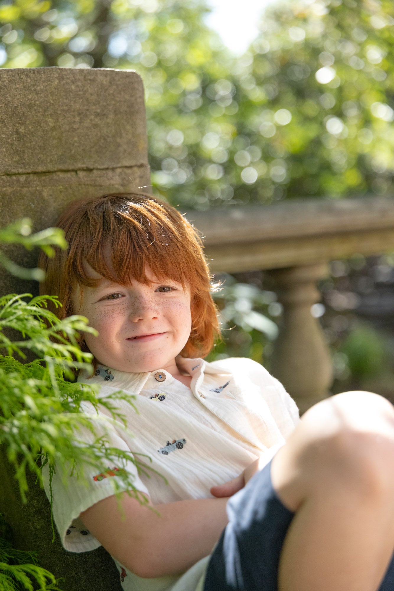 Child with red hair sitting outdoors near a stone column and greenery