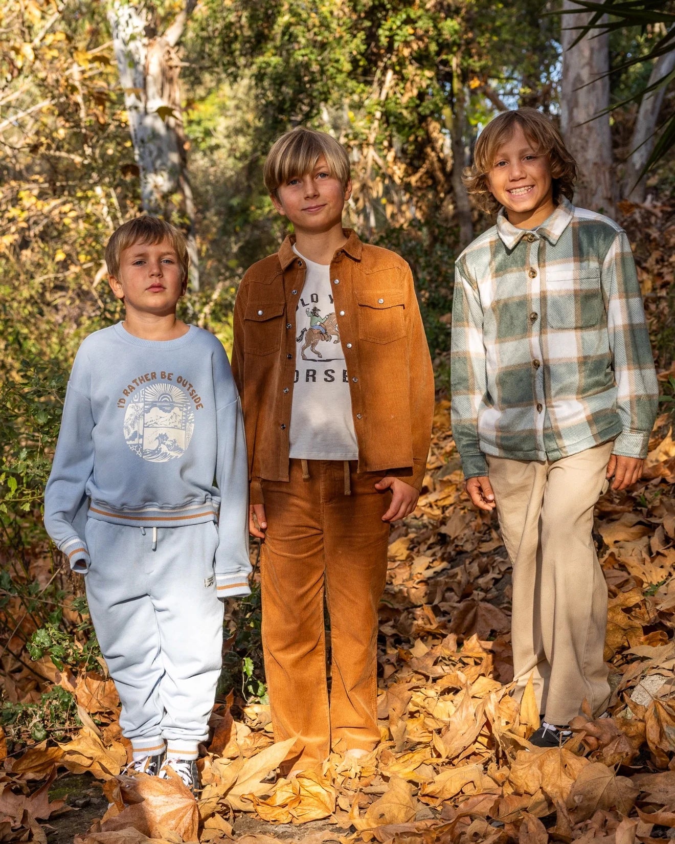 Three boys standing outdoors in a wooded area covered with fallen leaves, wearing different coordinated outfits from the brand Feather 4 Arrow.
