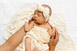 A sleeping newborn baby wrapped in a cream blanket with tiny red cherry prints, wearing a matching cream bow headband and swaddle glove, held by an adult's hands on a white textured blanket.