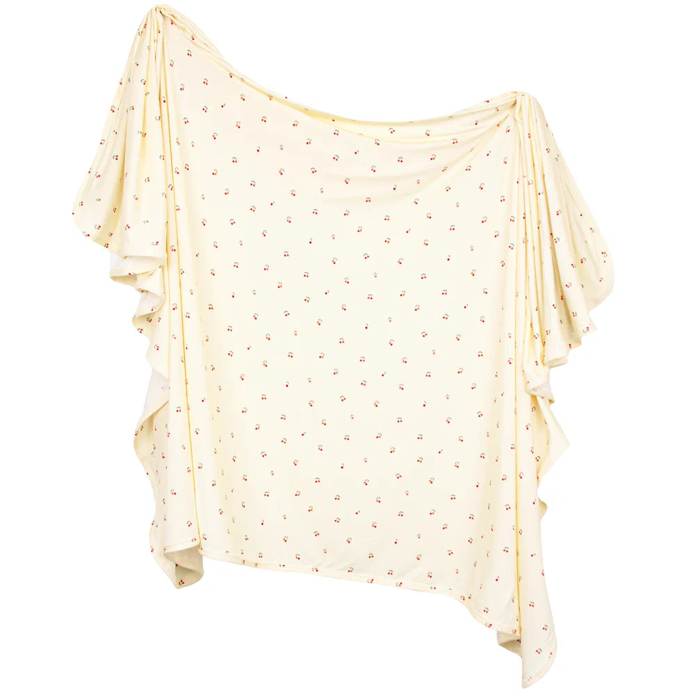 A cream blanket with a tiny red cherry print, featuring a draped silhouette, shown against a plain white background.