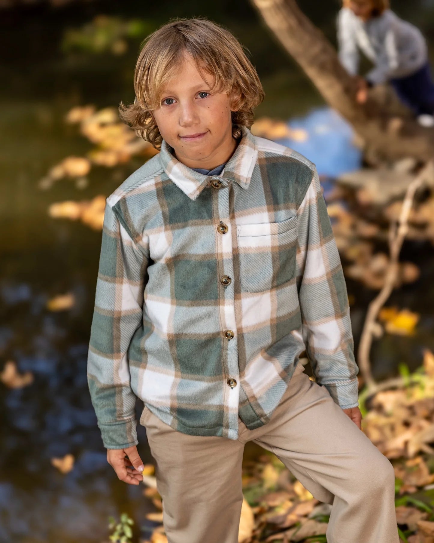 A young boy wearing a long-sleeve plaid button-up shirt in sage green, beige, and white, paired with khaki pants, standing outdoors near a stream with autumn leaves.