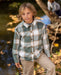 A young boy wearing a long-sleeve plaid button-up shirt in sage green, beige, and white, paired with khaki pants, standing outdoors near a stream with autumn leaves.