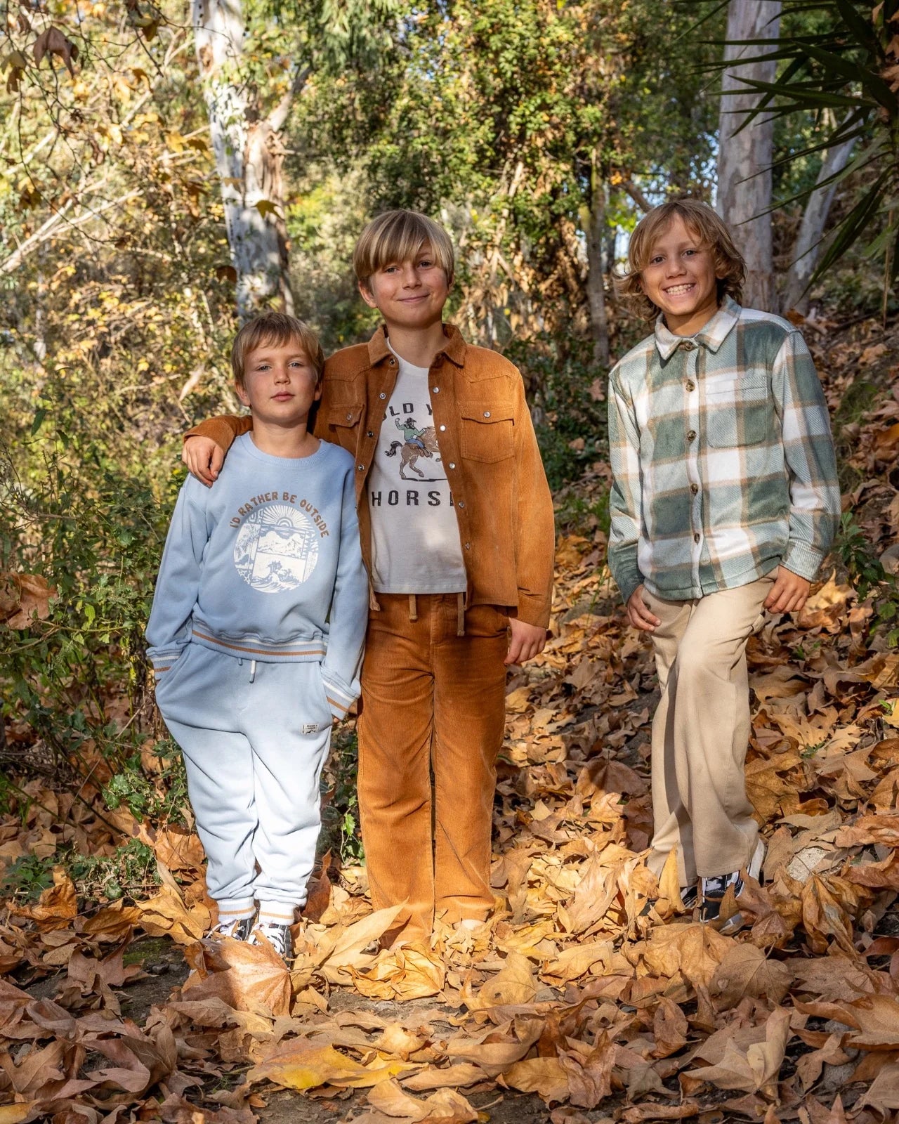 Three boys standing together outdoors on a ground covered in fallen leaves, wearing casual autumn clothing. The boy in the center wears a brown jacket and matching brown pants over a graphic T-shirt, the boy on the left wears a light blue sweatshirt and sweatpants, and the boy on the right wears a green plaid shirt and beige pants.