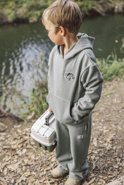 A young boy wearing a matching green hooded sweatshirt and sweatpants stands outdoors near a body of water, holding a white and gray fishing tackle box. The hoodie has a small embroidered fisherman logo on the left chest.