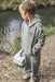 A young boy wearing a matching green hooded sweatshirt and sweatpants stands outdoors near a body of water, holding a white and gray fishing tackle box. The hoodie has a small embroidered fisherman logo on the left chest.