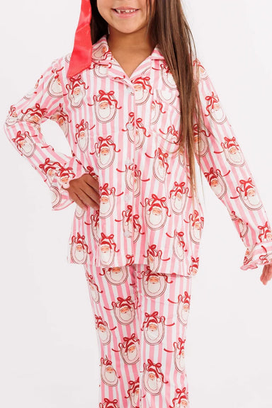 A young girl stands against a plain white studio background wearing a pink pajama set with a red bow in her hair. The pajamas have a white base with a repeating red and pink santa print.