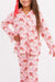 A young girl stands against a plain white studio background wearing a pink pajama set with a red bow in her hair. The pajamas have a white base with a repeating red and pink santa print.