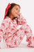 A young girl sitting against a plain white studio background wearing a pink pajama set with a red bow in her hair. The pajamas have a white base with a repeating red and pink santa print.