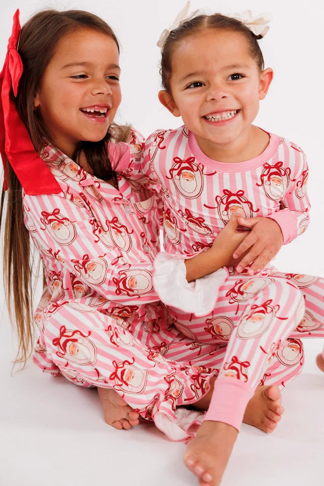 Two young girls sitting together wearing matching pink and white striped pajamas with a Santa Claus print, against a plain white background.