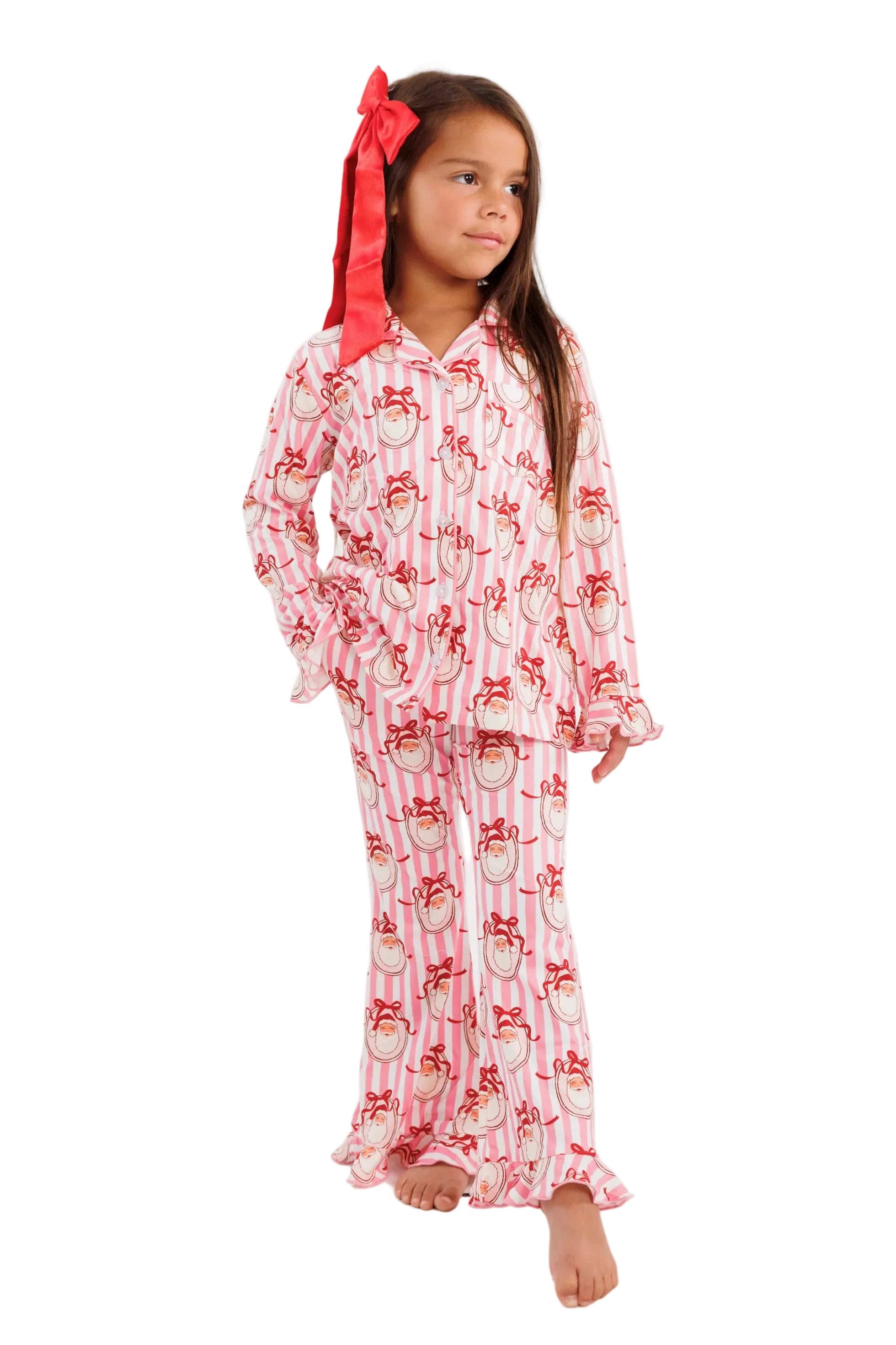 A young girl stands against a plain white studio background wearing a pink pajama set with a red bow in her hair. The pajamas have a white base with a repeating red and pink santa print.