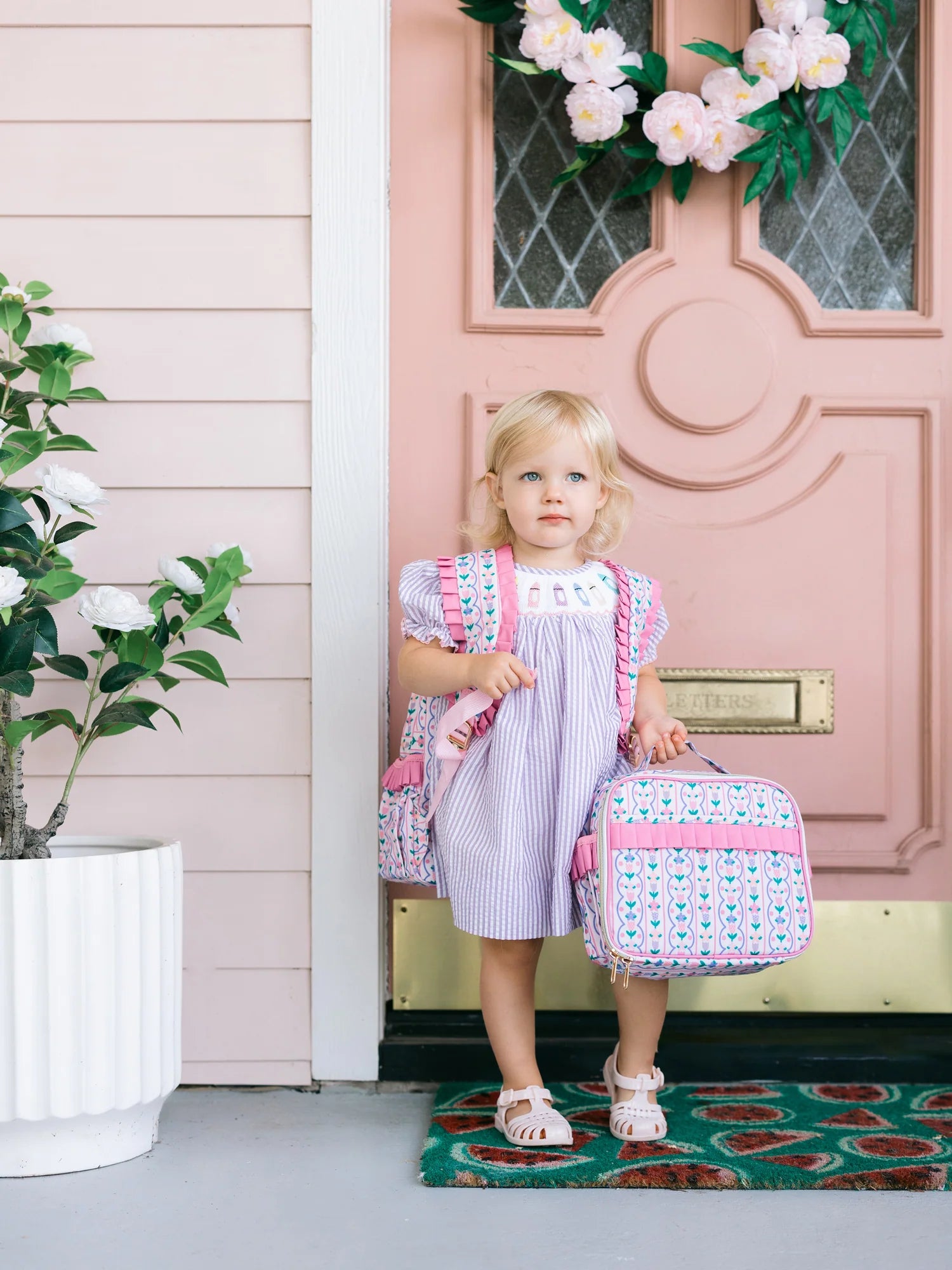 Young girl holding a colorful lunchbox in front of a pink door with floral decorations