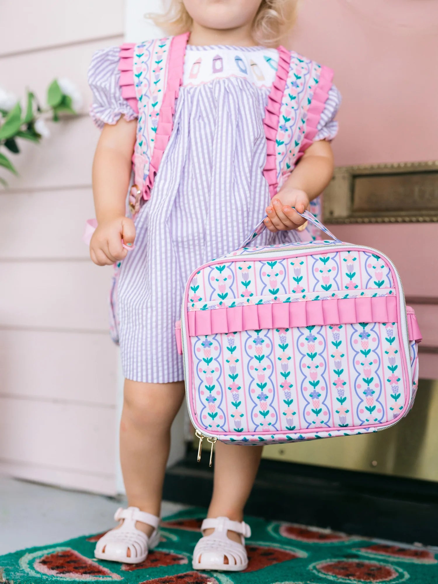 Child holding a patterned lunchbox with pink straps in a home setting