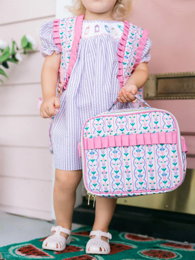 Child holding a patterned lunchbox with pink straps in a home setting