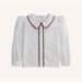 A white long-sleeve blouse for toddlers, featuring a Peter Pan collar and front placket trimmed with red scalloped fabric, shown against a light cream background.