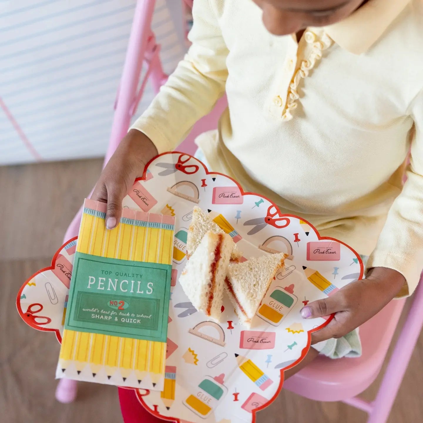 Person holding a box of pencils and a plate with sandwiches on a pink chair.