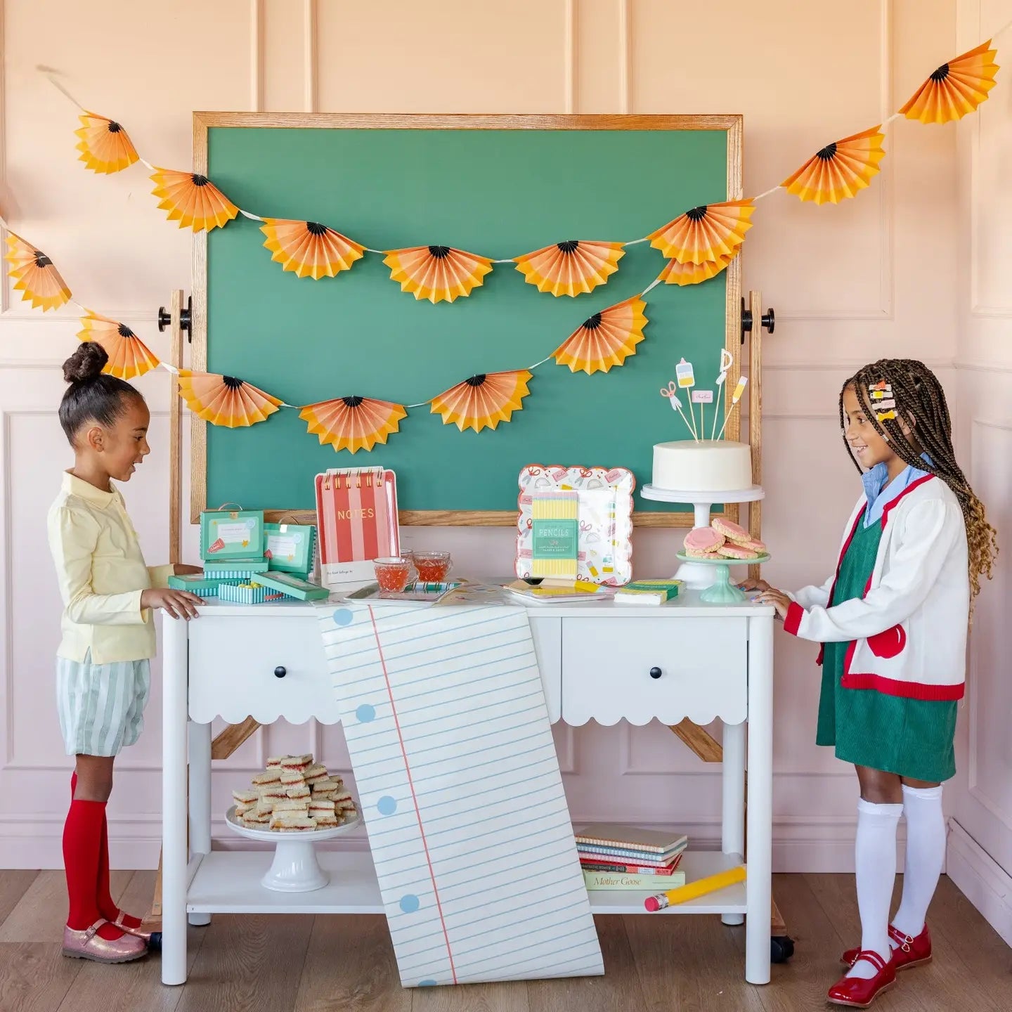 Two children standing in front of a table with decorative items and a green chalkboard in the background.
