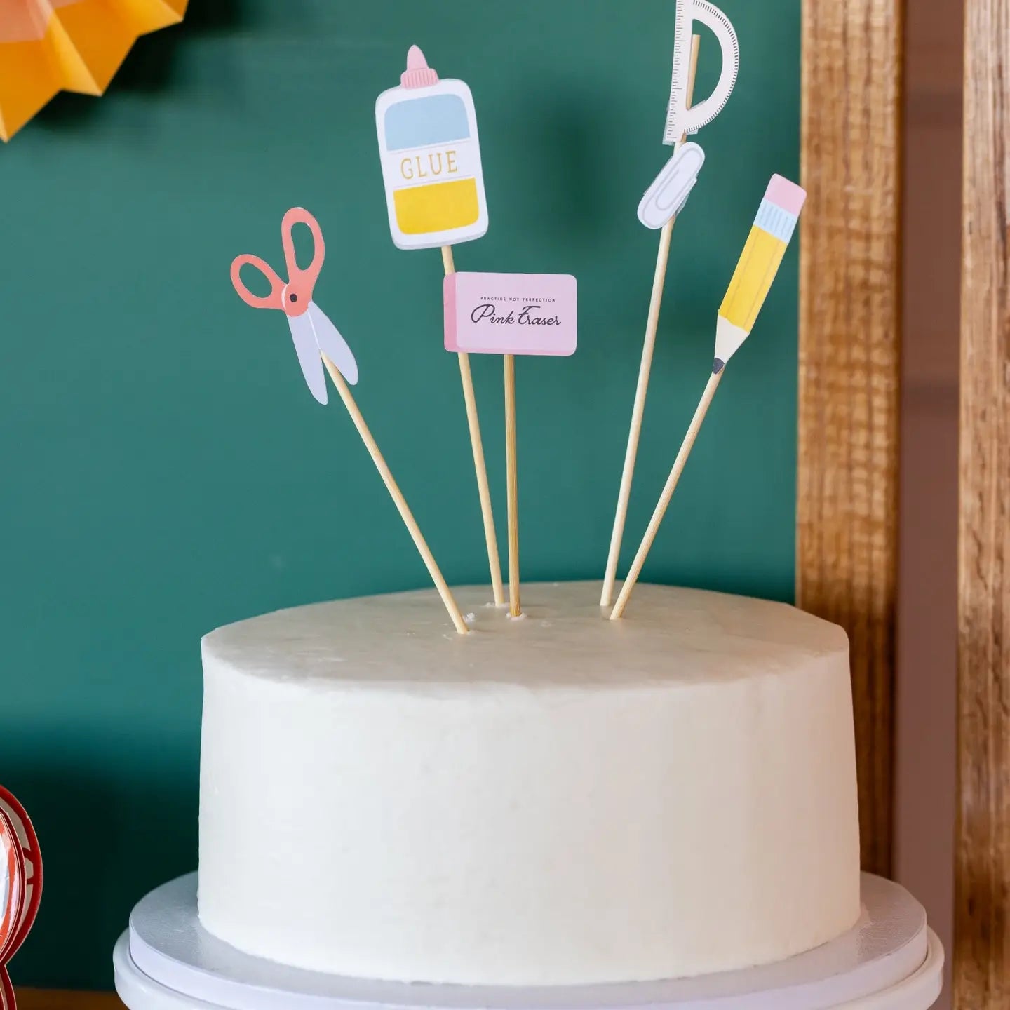 White cake with decorative toppers resembling school supplies against a green background