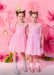 Two young girls in pink dresses standing against a pink floral background