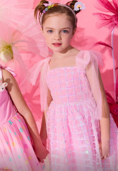 Young girl in a pink dress with floral headband standing against a pink background