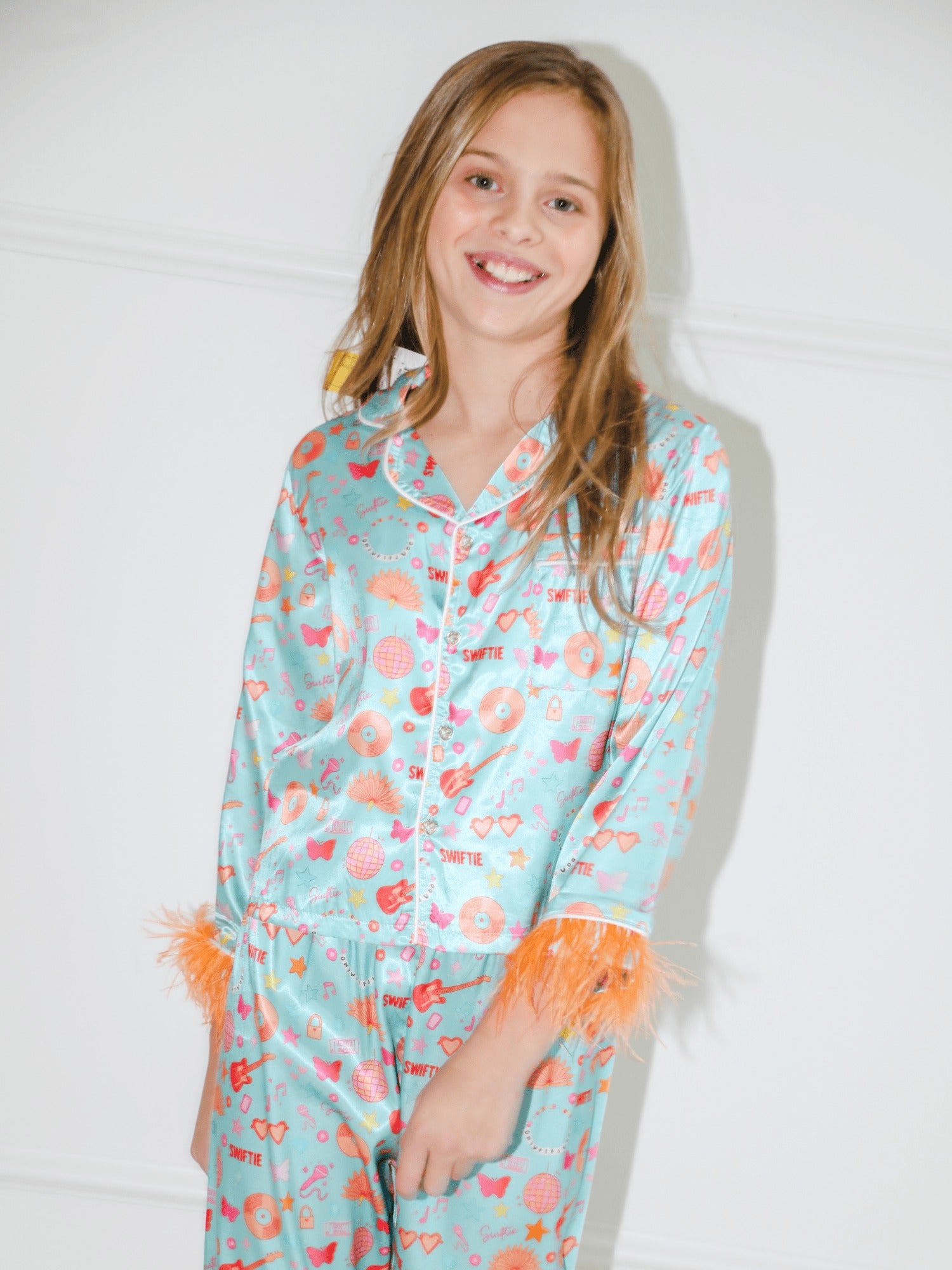 A young girl smiling while wearing a light blue satin pajama set with a colorful heart and star pattern, orange feather trim at the cuffs, and orange feather accents at the waist.