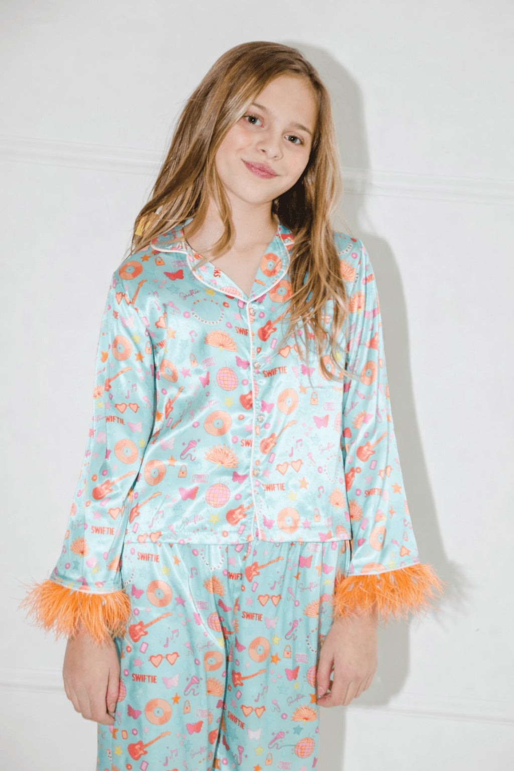 A young girl wearing a two-piece satin pajama set with a light blue base, orange and "Swifty" patterns, and orange feather trim on the cuffs, standing against a white wall.