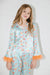 A young girl wearing a two-piece satin pajama set with a light blue base, orange and "Swifty" patterns, and orange feather trim on the cuffs, standing against a white wall.