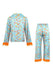 A two-piece pajama set featuring a long-sleeve button-up shirt and matching pants in light blue satin with a colorful pattern of sunglasses, guitars, and flowers, finished with bright orange fringe trim at the cuffs and hem.