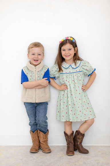 Two children modeling coordinated outfits against a plain white wall. The boy wears a beige vest over a blue shirt with jeans and brown cowboy boots.