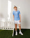 Child in a blue striped polo shirt and white shorts standing in a room with a white picket fence.