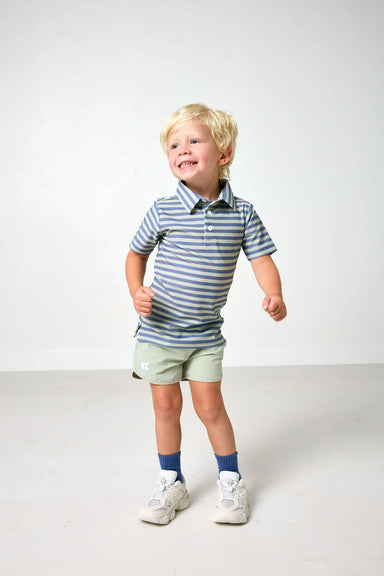 A young blonde boy wearing a blue and green striped polo shirt, light green shorts, blue socks, and white sneakers, standing against a plain light gray studio background.