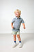 A young blonde boy wearing a blue and green striped polo shirt, light green shorts, blue socks, and white sneakers, standing against a plain light gray studio background.