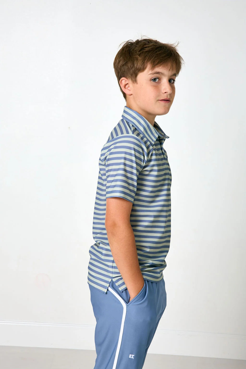 A young boy standing in profile wearing a blue and green striped short-sleeve polo shirt and matching blue track pants with a white side stripe, against a plain white wall.