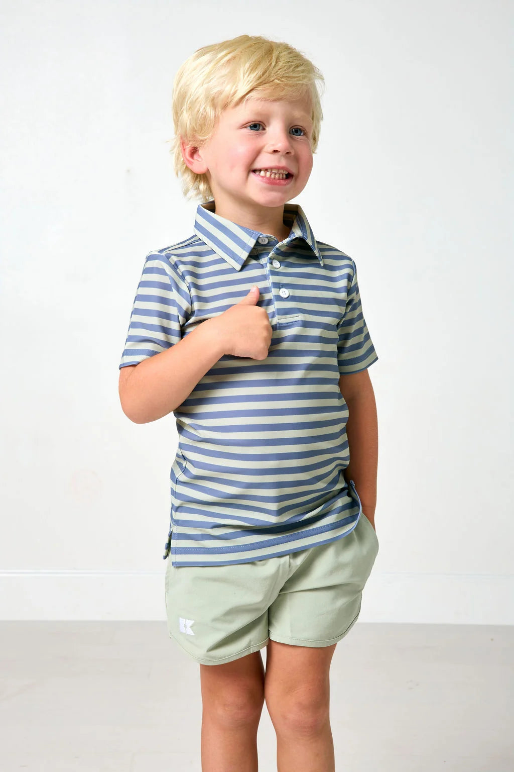 A young blonde boy smiling and giving a thumbs-up, wearing a blue and green striped short-sleeve polo shirt and light sage green shorts, standing against a plain white studio background.