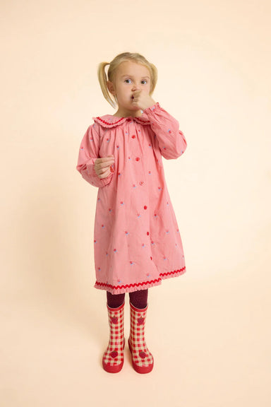 A young girl with blonde pigtails stands against a solid pale peach background wearing a pink long-sleeve nightgown with small red and blue polka dots, dark red tights, and red-and-white checkered rain boots.