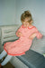 A young girl with blonde hair in a ponytail sits on a striped cushioned bench, wearing a pink long-sleeve dress with red trim and white tights.