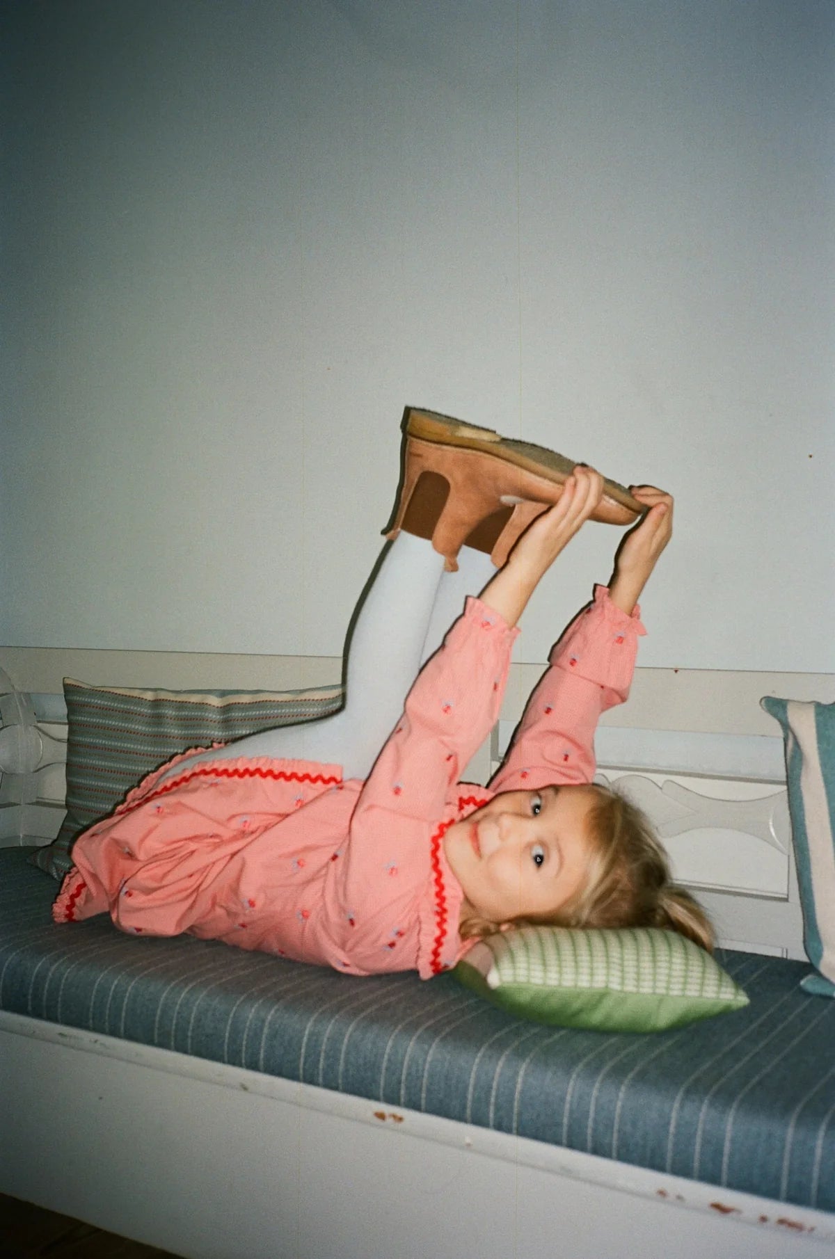 A young girl lies on a bed stretching her leg upward while wearing a pink long-sleeve top with small red dots and red trim, white tights, and brown wedge sandals.