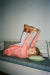 A young girl lies on a bed stretching her leg upward while wearing a pink long-sleeve top with small red dots and red trim, white tights, and brown wedge sandals.