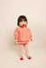 A toddler standing against a plain light beige studio background, wearing a peach-pink long-sleeve top with small embroidered details and red trim, matching pink leggings, and red-and-white socks. The child has dark hair and is holding a red pacifier.