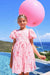 A young girl stands by a swimming pool holding a large pink balloon, wearing a light pink dress with darker pink bow and ribbon patterns. The background features a clear blue sky, ocean, and green hills.
