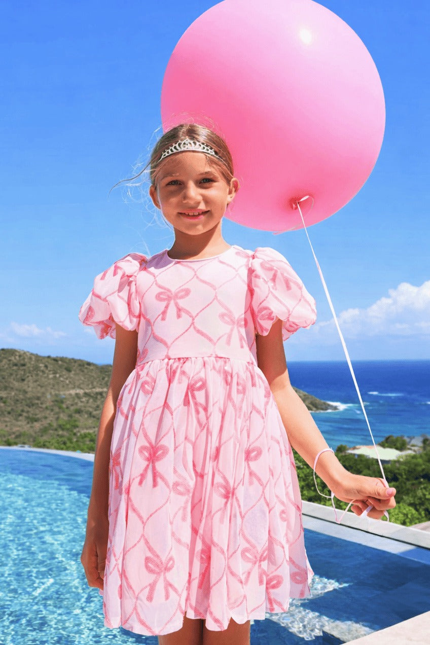 A young girl stands by a swimming pool holding a large pink balloon, wearing a light pink dress with darker pink bow and ribbon patterns. The background features a clear blue sky, ocean, and green hills.