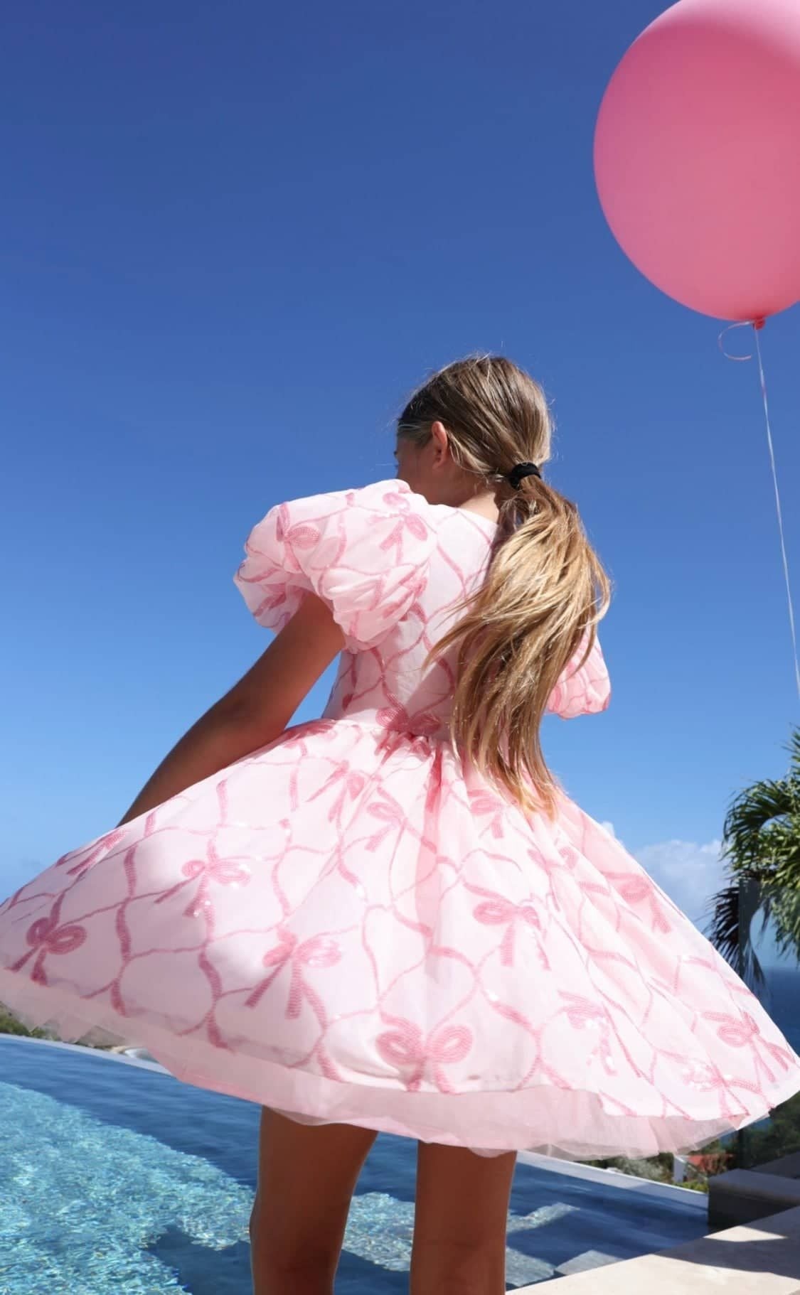 Person in a pink dress with floral pattern holding a pink balloon by a pool.