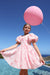 A young girl wearing a pink and white dress with a bow pattern, standing in front of a blue swimming pool and sky while holding a large pink balloon.