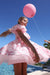 A young girl wearing a pink dress with a bow pattern twirls outdoors near a swimming pool, holding a large pink balloon. A hand on the right holds the balloon string.