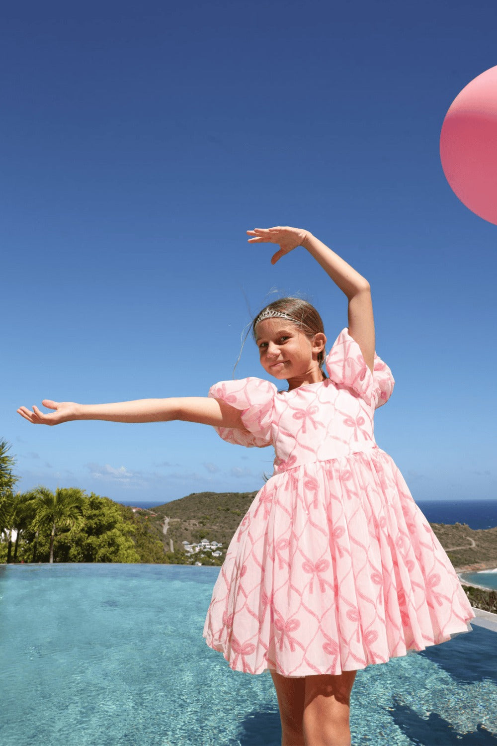 A young girl wearing a pink patterned dress stands in a swimming pool with a clear blue sky and distant hills in the background. A pink balloon is partially visible in the top right corner.