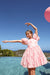 A young girl wearing a pink patterned dress stands in a swimming pool with a clear blue sky and distant hills in the background. A pink balloon is partially visible in the top right corner.