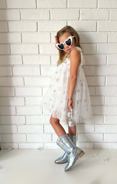 Child wearing a white dress with ruffles and silver rain boots against a white brick wall.